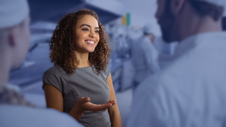 A smiling woman with curly hair talking to two chefs in a kitchen