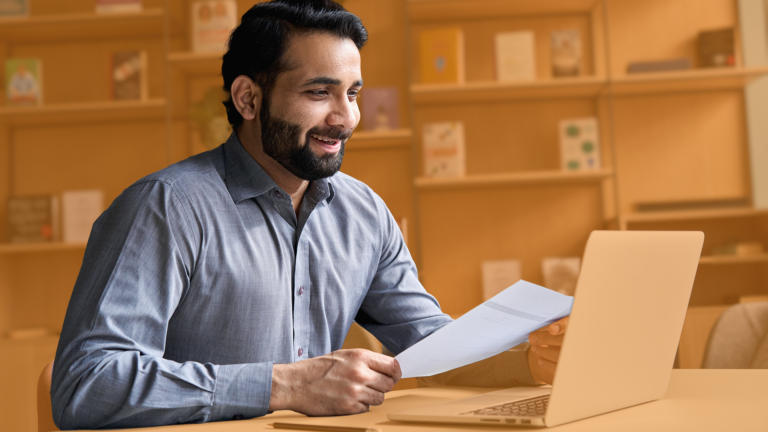 A smiling man at home working on a laptop holding a piece of paper