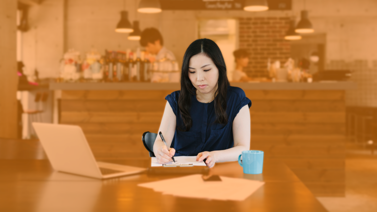 A girl writing on a paper with mug and laptop on the table